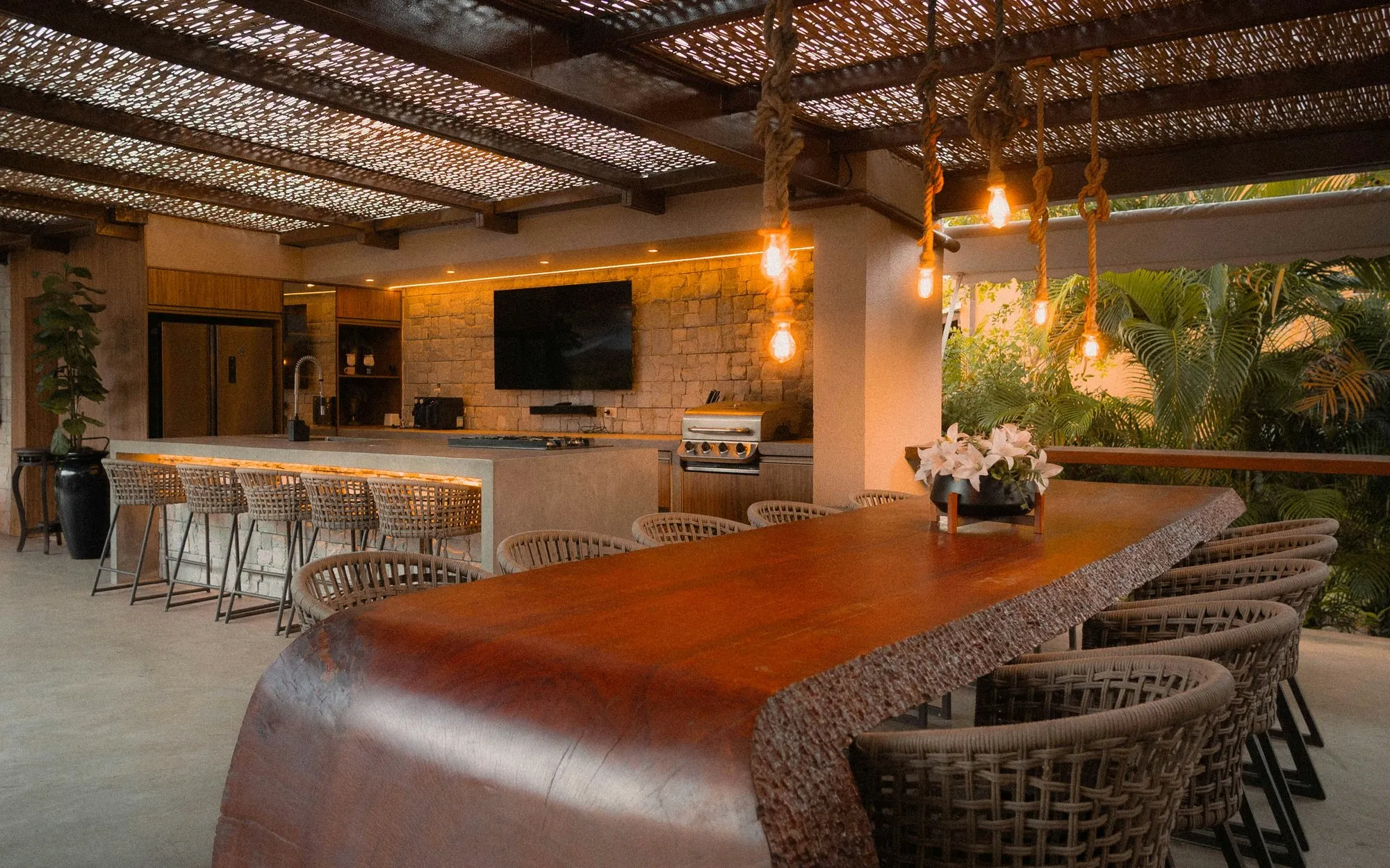 Überdachte Outdoorküche mit Kochzeile, Theke und langer Tafel unter einer Pergola aus Schilf